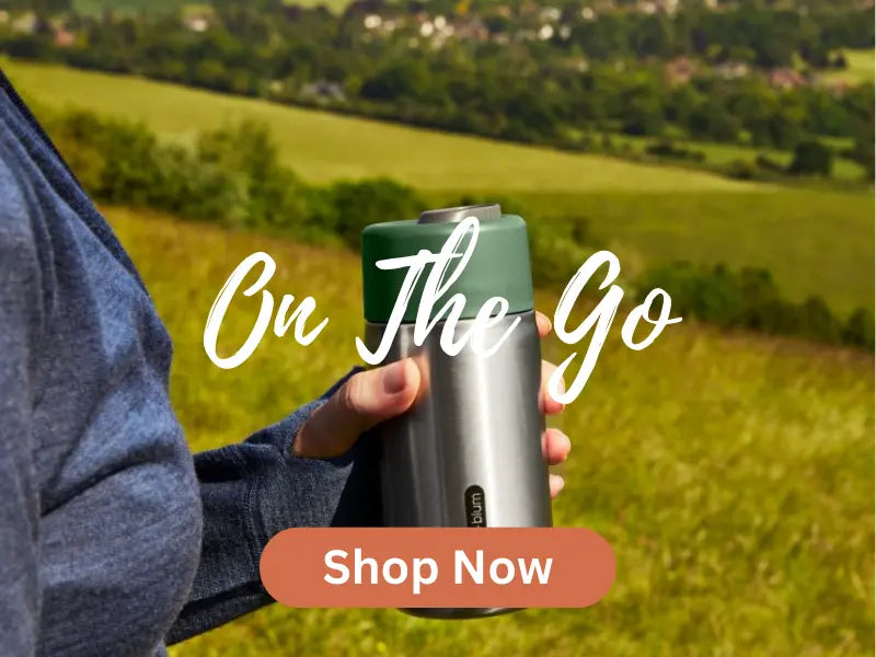 Reusable stainless steel travel mug with a green lid, held outdoors in a scenic countryside setting. Perfect for eco-friendly hydration on the go. Shop now for sustainable travel essentials.